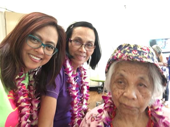 Three women in floral leis share smiles and laughter at a joyful celebration.