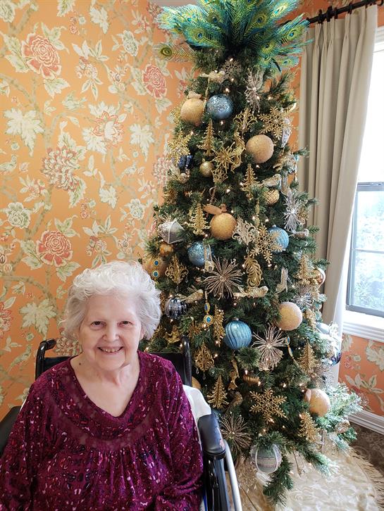 An elderly woman sits happily in front of a beautifully decorated Christmas tree in a warm room.