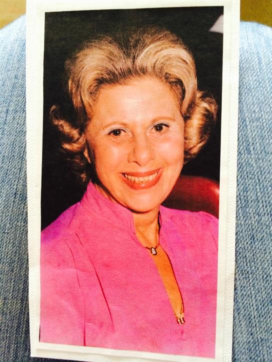 A woman with curly hair in a pink blouse smiles warmly at an event surrounded by friends.
