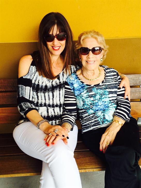 Two women, one young and one older, sit on a bench smiling and wearing sunglasses on a sunny day.