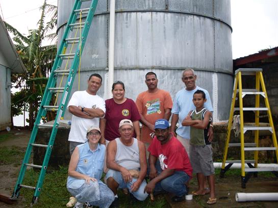 Seven individuals pose smiling by a large water tank, showcasing teamwork in rural outreach.