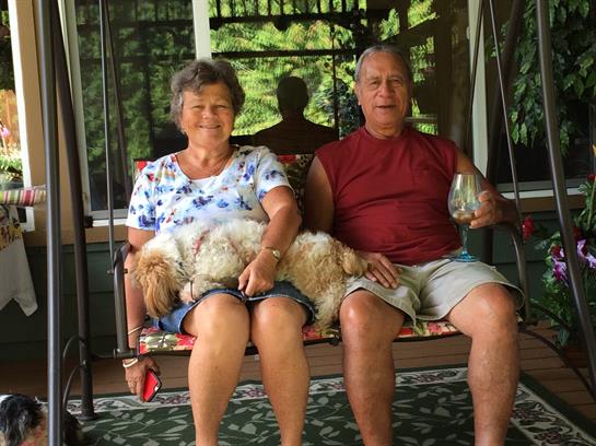 Couple sits on a swing on a porch, enjoying drinks while their dog rests on their laps.