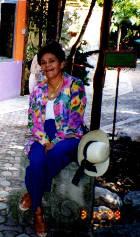 Woman in bright attire sits on a stone bench enjoying a sunny day in a lively location.