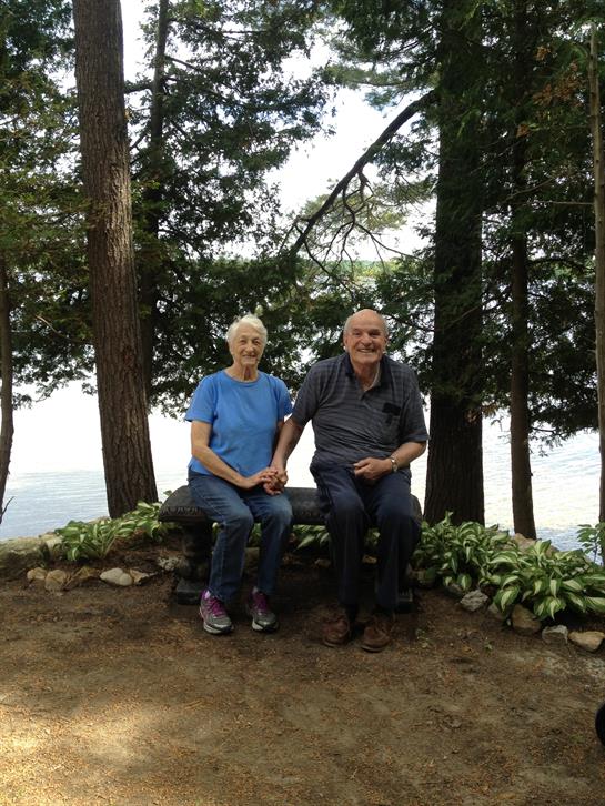 An elderly couple sits together holding hands on a bench by the lake, surrounded by lush greenery.