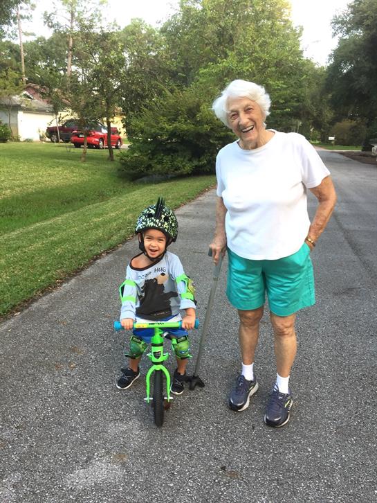 A young boy enjoys riding his bicycle while his grandmother smiles proudly beside him outdoors.