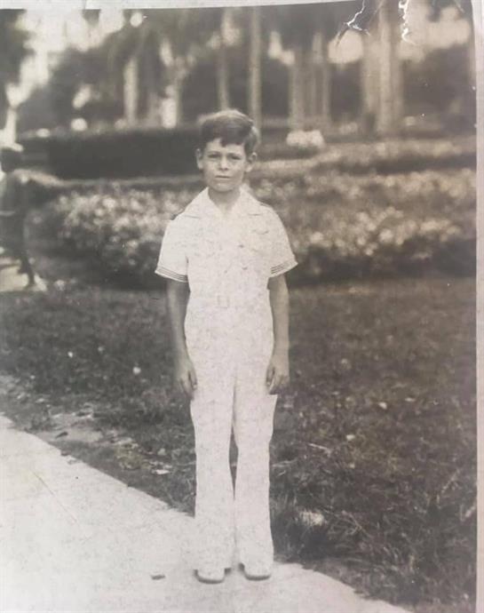 A boy wearing a white outfit stands in a well-kept garden filled with flowers.