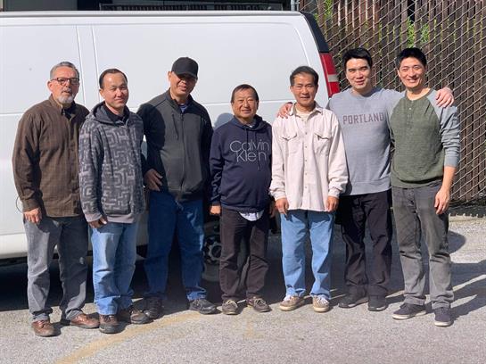 Eight men smile together outdoors, enjoying a warm, sunny day near a white van.