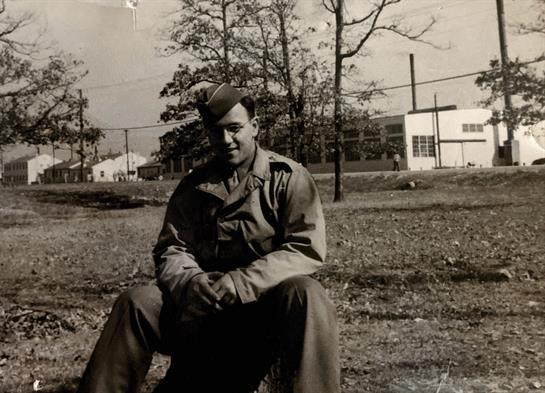 A soldier relaxes on the grass in military attire, enjoying a sunny day near barracks.