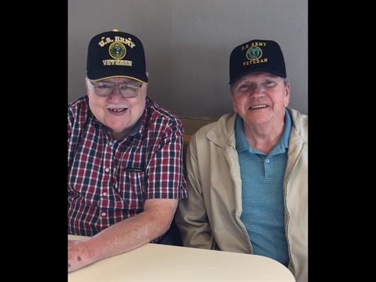 Two smiling elderly men sit at a table, sharing stories and laughter at a local community center.