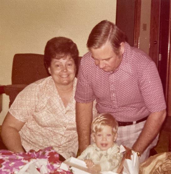 A couple watches with joy as their young child unwraps gifts in a warm living room.
