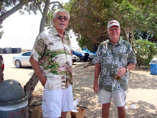 Two men chat and relax in casual attire under trees by the beach, enjoying drinks on a sunny day.
