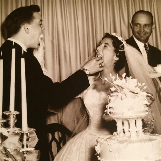 A couple joyfully cuts their wedding cake, surrounded by celebrating guests.