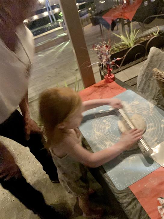 A child enthusiastically rolls out dough while an adult guides her in a vibrant outdoor setting.