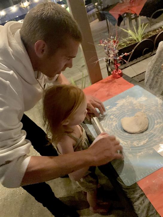 A father teaches his daughter to roll dough at a colorful table in their home kitchen.