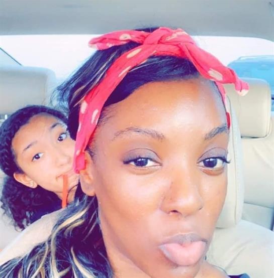 A mother playfully makes a face while her daughter poses behind her in the car.