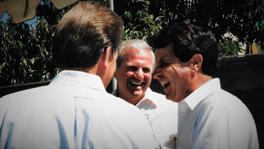 Three smiling men in white shirts laugh together outdoors, enjoying the sun.