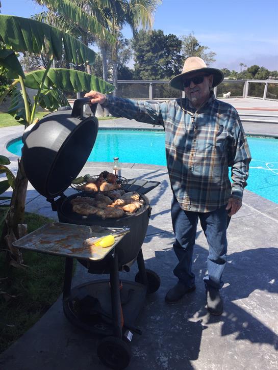 A man stands beside a grill, cooking chicken and steak outdoors near a pool on a sunny day.