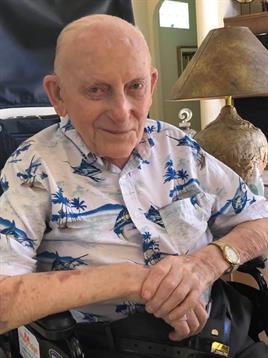 An elderly man sits comfortably in a cozy living room, wearing a Hawaiian shirt and smiling gently.