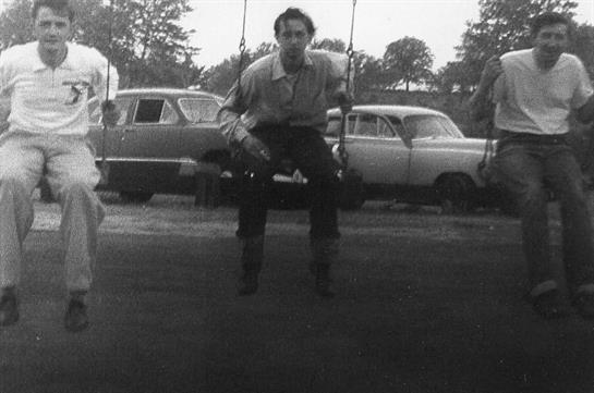 A young man enjoys swinging at a public park surrounded by classic cars on a bright day.