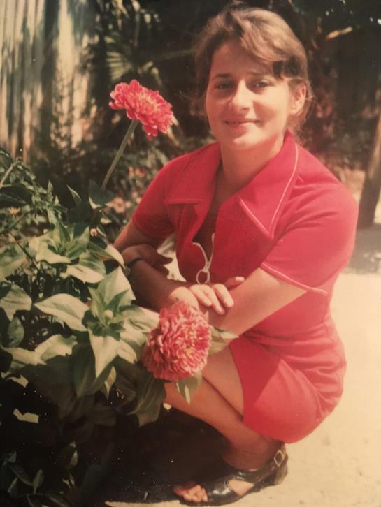 Woman smiles while kneeling beside colorful flowers in a bright garden setting.