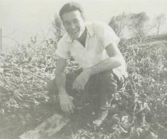Young man kneels in a field, smiling broadly while holding a piece of paper in a rural setting.