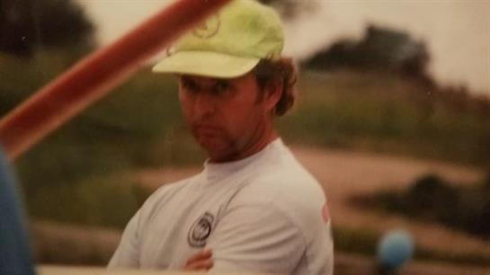 A man in a green cap stands outdoors with arms crossed, looking serious during a sunset.