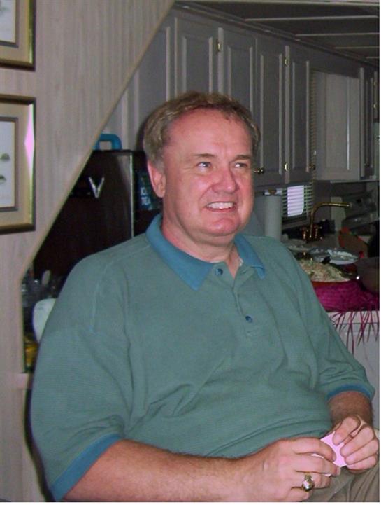 A man in a green sweater smiles in a cozy kitchen with family and friends around.