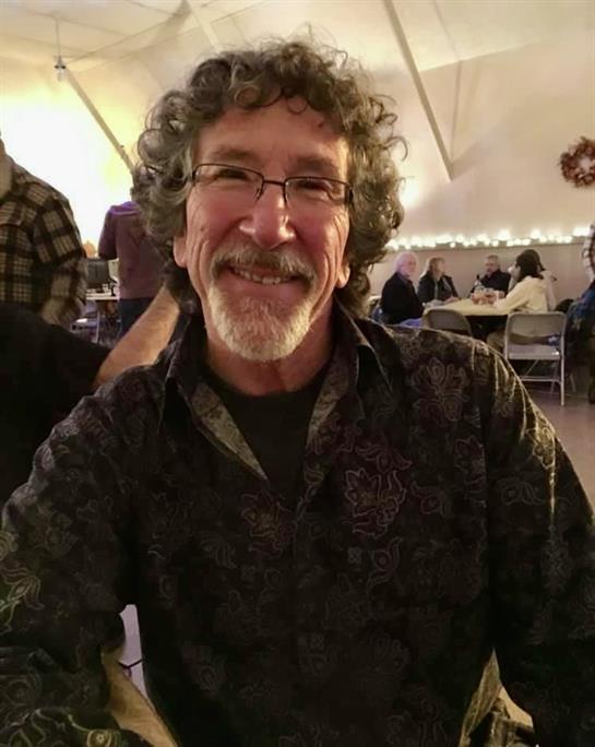 A cheerful man with curly hair and glasses poses for a picture while seated at a festive gathering.