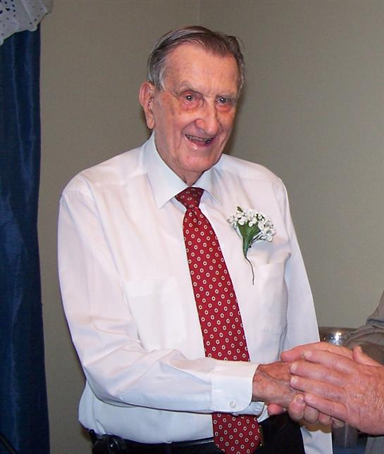 Elderly man with a bouquet on his shirt shakes hands with another individual inside a room.