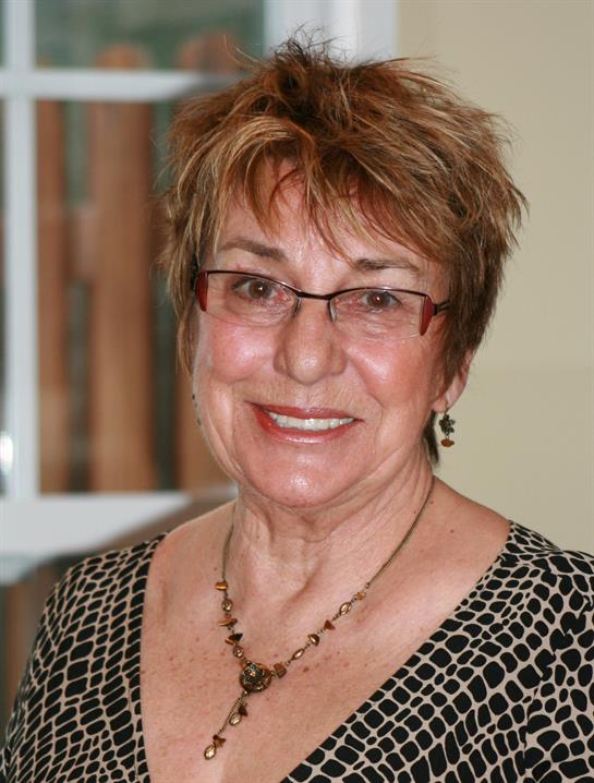 A cheerful woman with glasses is smiling warmly in a comfortable room filled with soft lighting.