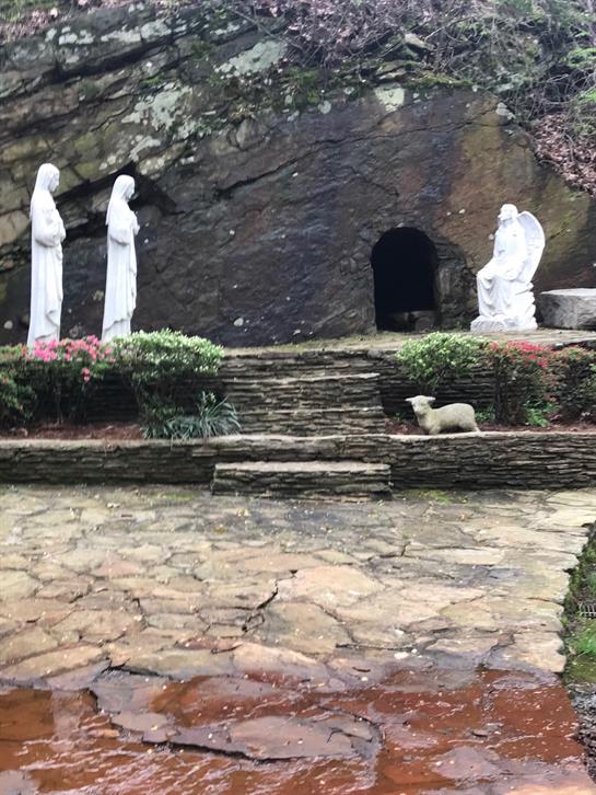 Religious statues are arranged in a tranquil garden setting near a rocky cave entrance.