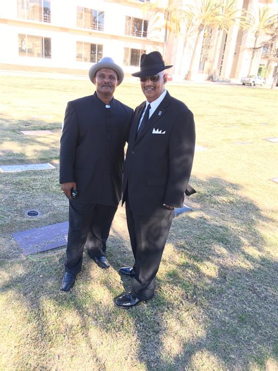 Two well-dressed men stand together smiling on a grassy area in a park.