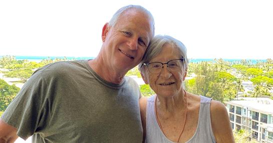 Two family members smile for a photo on a balcony, with the ocean in the background.