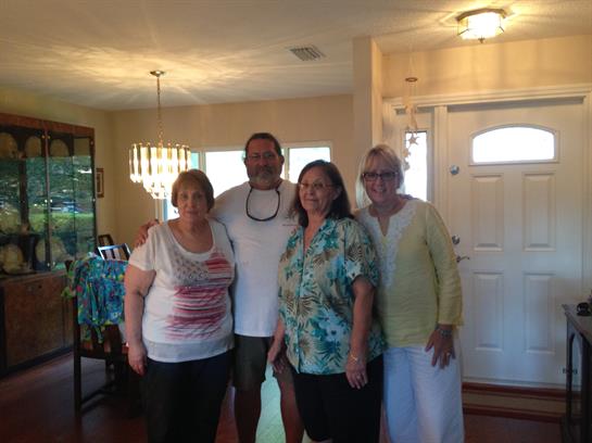 Four friends stand together, smiling warmly inside a cozy living room filled with light.