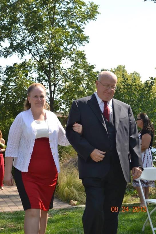 Two individuals walk together at an outdoor wedding ceremony, dressed elegantly for the occasion.
