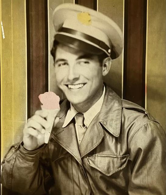 A smiling young man in a military uniform holds an ice cream cone, seated indoors.