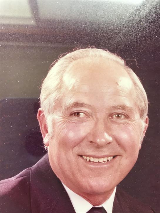 An elderly man with gray hair smiles warmly while wearing a formal outfit in a bright interior.