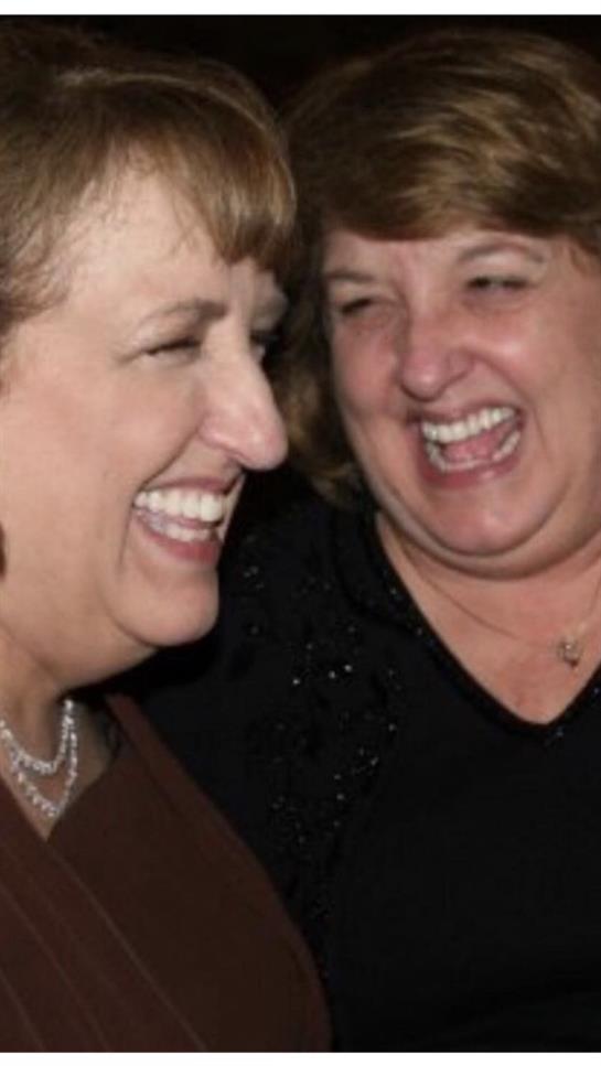 Two women are laughing heartily while engaging warmly at a social gathering, showcasing friendship.