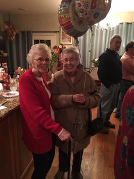 Two elderly women stand together, sharing smiles and conversation during a cozy gathering.