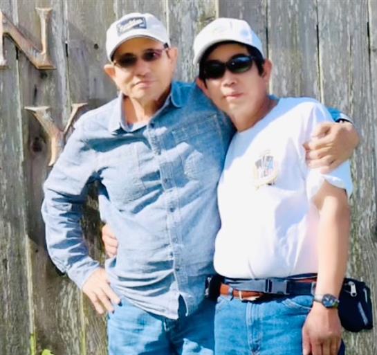 Two men stand smiling by a rustic wooden fence in bright sunlight.