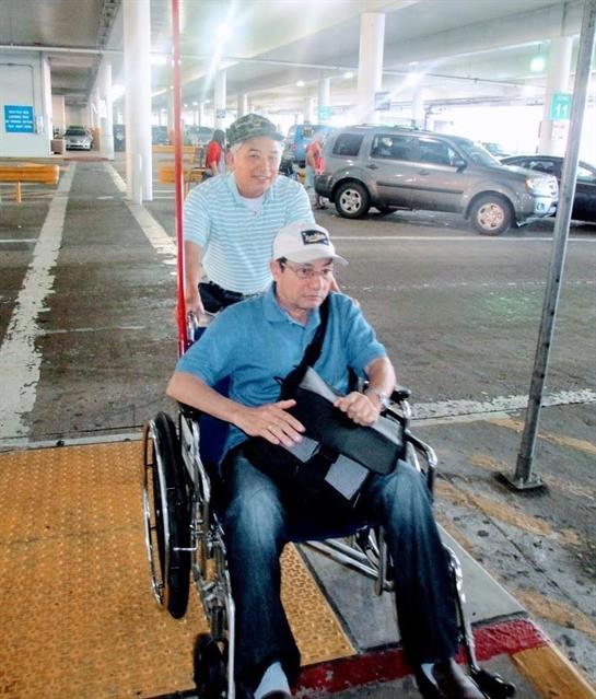 Two individuals are navigating a parking lot; one uses a wheelchair while being helped by another.