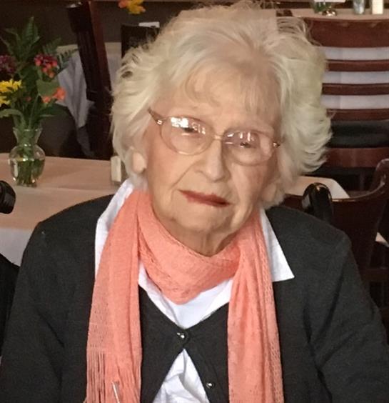 An elderly woman sits at a restaurant table, wearing a warm scarf and glasses, smiling softly.