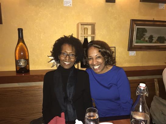 Two women smile joyfully while seated at a wooden table adorned with drinks in a restaurant.