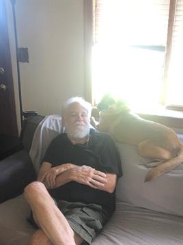 An elderly man sits back on a couch while a dog playfully interacts with him in a warm setting.