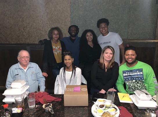A diverse group gathers around a table for a celebration, enjoying food and each other’s company.