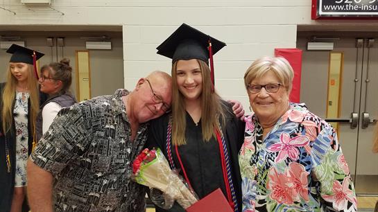 A young graduate stands with her grandparents, celebrating her graduation with flowers and joy.