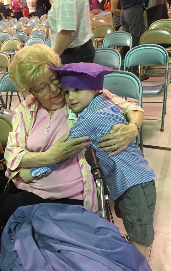 A child hugs an elderly woman at a community gathering. They sit in a large hall with chairs around.