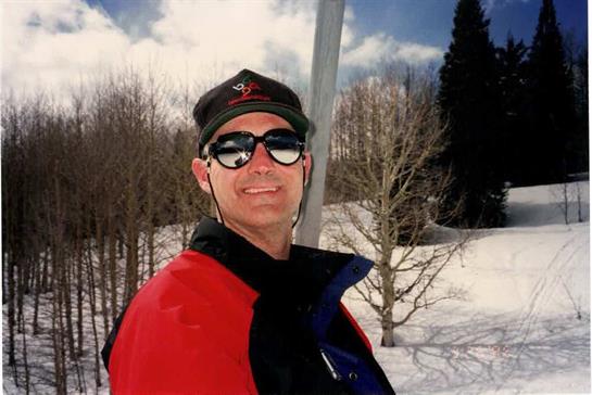 A man smiles while wearing sunglasses, dressed for skiing in a snowy landscape.