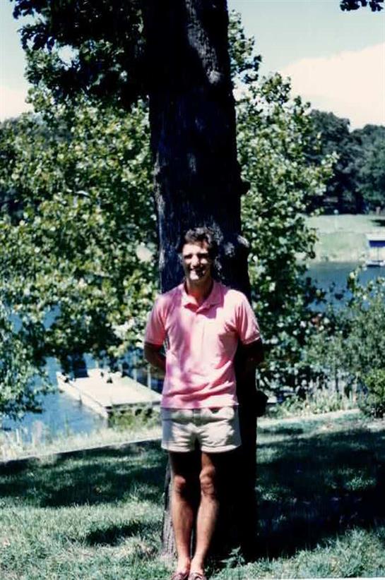A person poses next to a tree with a lake in the background, capturing a peaceful summer moment.