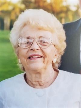 An elderly woman with light hair smiles warmly in a sunny park surrounded by nature.
